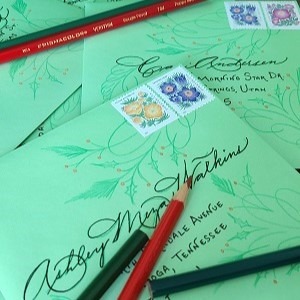 A pile of green envelopes with holly and pine flourishes and colored pencils used to make the artwork